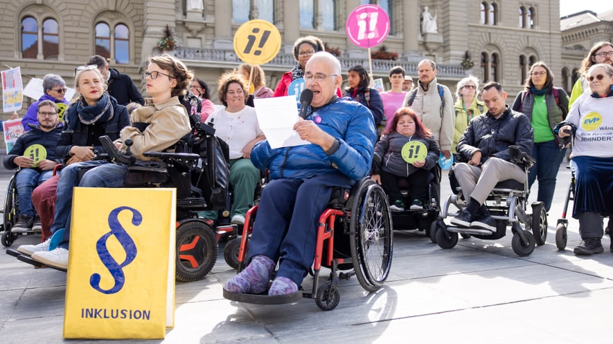 Eine Gruppe von Menschen, darunter viele im Rollstuhl, hält Plakate hoch und versammelt sich bei einer Protest-Veranstaltung vor dem Bundeshaus zum Thema Inklusion. Eine Person im Vordergrund liest einen Text vor.