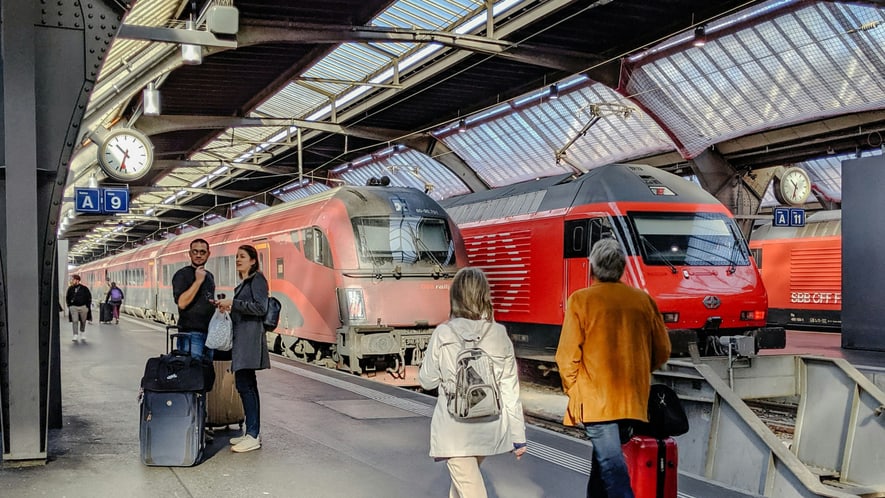 Two red trains are stopped at a covered station platform as several people with luggage walk and wait nearby.